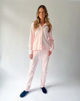 A woman modeling a light pink two-piece pajama set consisting of a long-sleeve button-up shirt and matching pants, paired with dark blue slippers, against a light gray studio background.