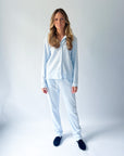 A woman modeling a light blue two-piece pajama set consisting of a long-sleeve button-up shirt and matching pants, paired with dark navy slippers, against a light gray studio background.