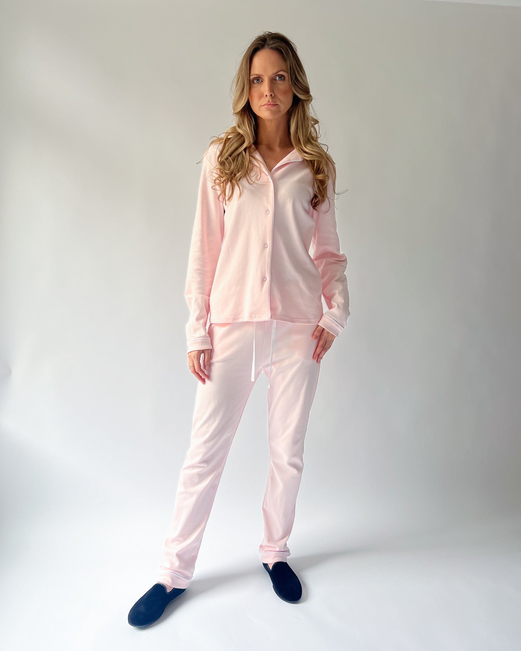 A woman modeling a light pink two-piece pajama set consisting of a long-sleeve button-up shirt and matching pants, paired with dark blue slippers, against a light gray studio background.
