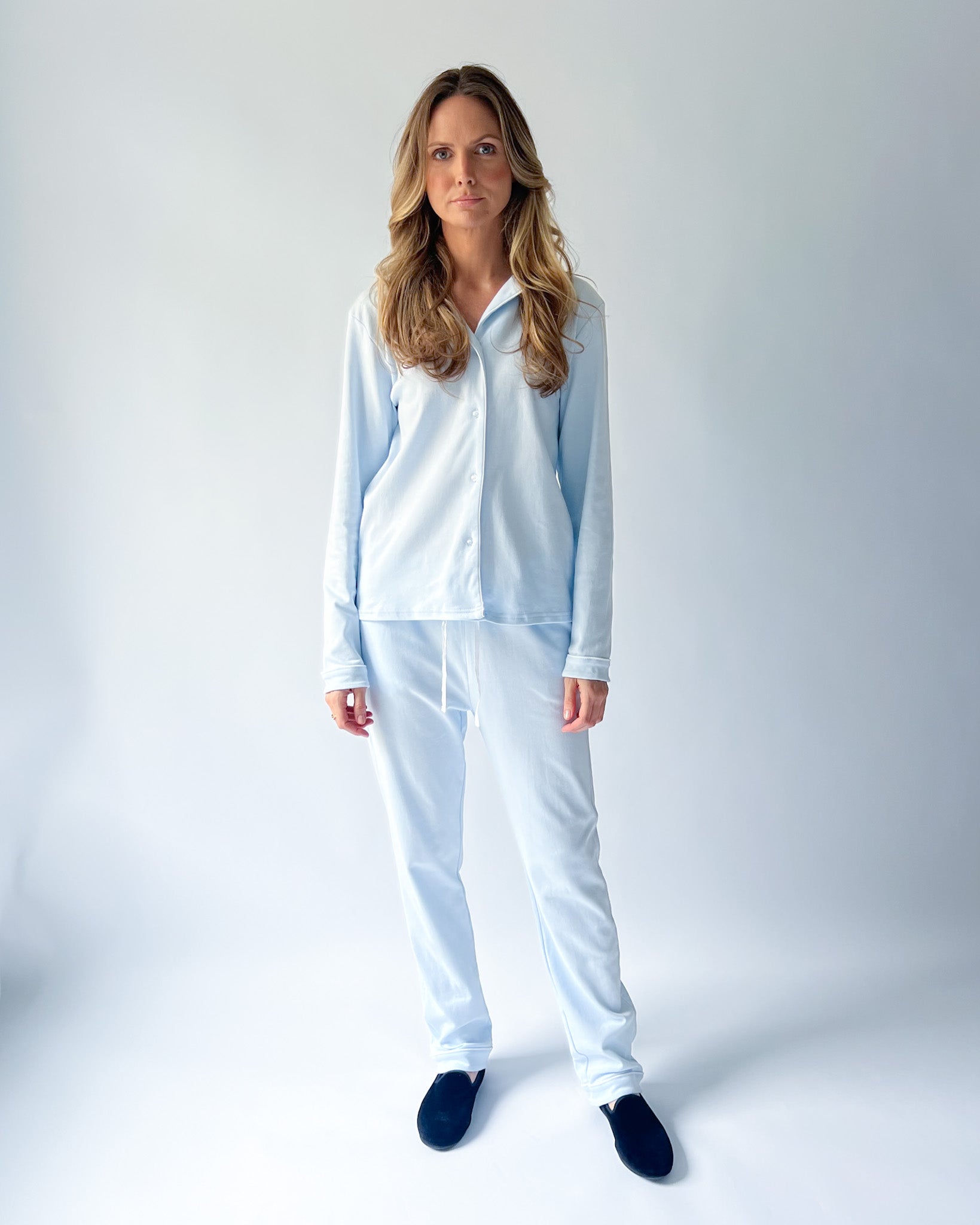 A woman modeling a light blue two-piece pajama set consisting of a long-sleeve button-up shirt and matching pants, paired with dark navy slippers, against a light gray studio background.